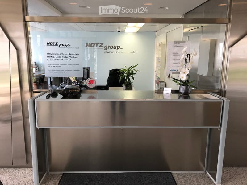 Reception desk with Notz group branding, plant, and orchid. Glass walls and doors. Business hours and contact information displayed.