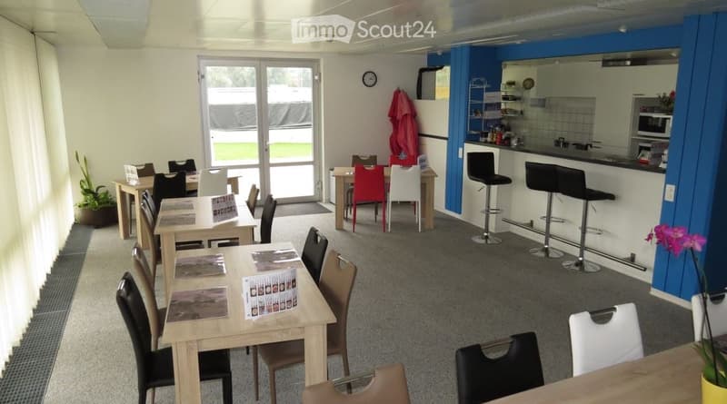 The image shows an open-plan living space with a dining area, kitchen, and lounge area. The space has large windows, a blue accent wall, and various furniture and decor items. The kitchen area is visible and appears to have a dishwasher.