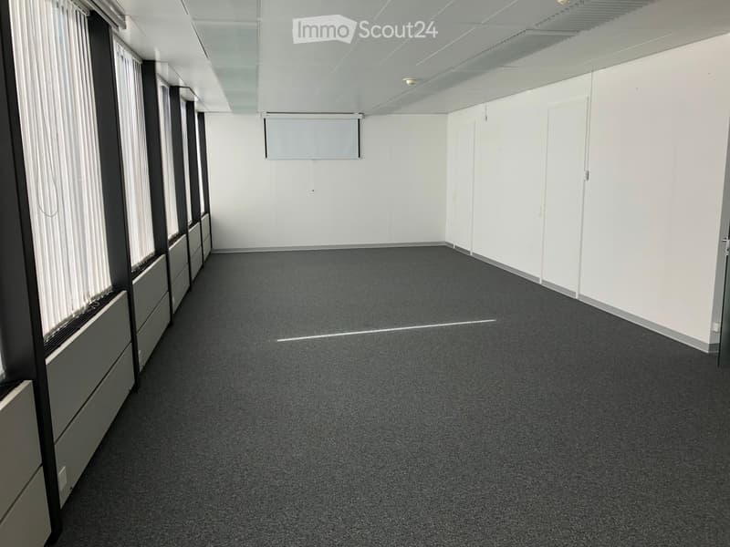 Large open room with gray carpeted floor, white walls, and a projector screen mounted on the wall. The room appears to be an office or meeting space with ample natural light from the windows.