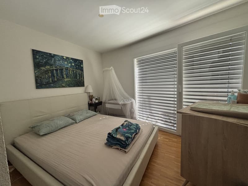A well-lit bedroom with a large bed, pillows, a nightstand with a lamp, a painting on the wall, and a small table with a mirror.