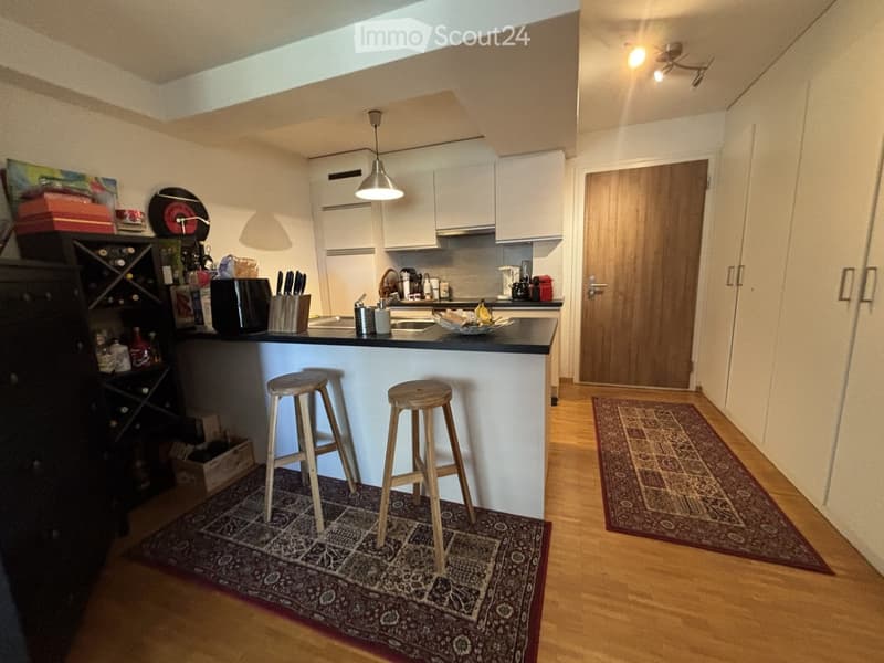Modern kitchen with white cabinets, bar counter, two wooden stools, wine rack, and various appliances.