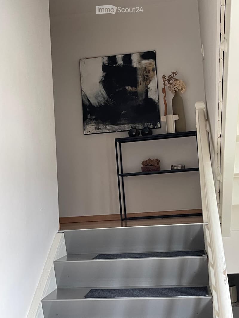Modern hallway with a staircase featuring metallic steps and a sleek black shelf on the wall holding decorative items, including a vase with flowers and a framed artwork.