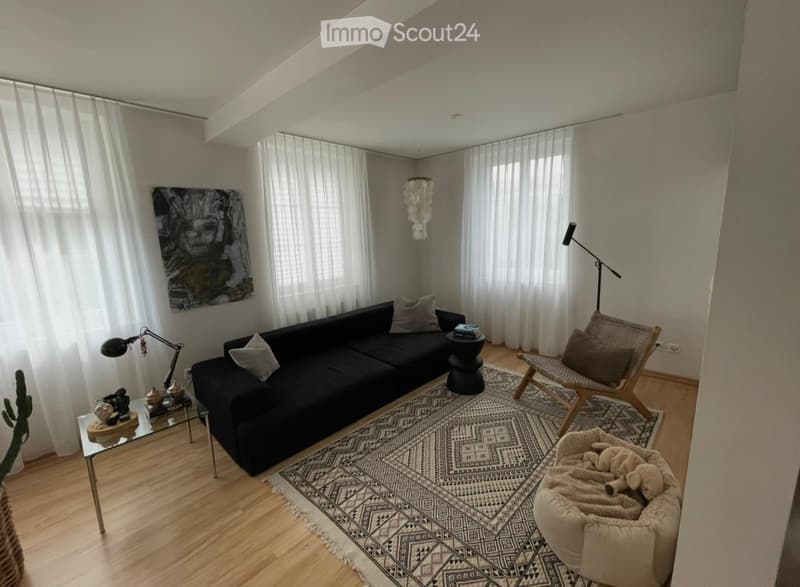 Modern living room with a black sectional sofa, wooden floor, patterned rug, floor lamp, and decorative items on a glass table.