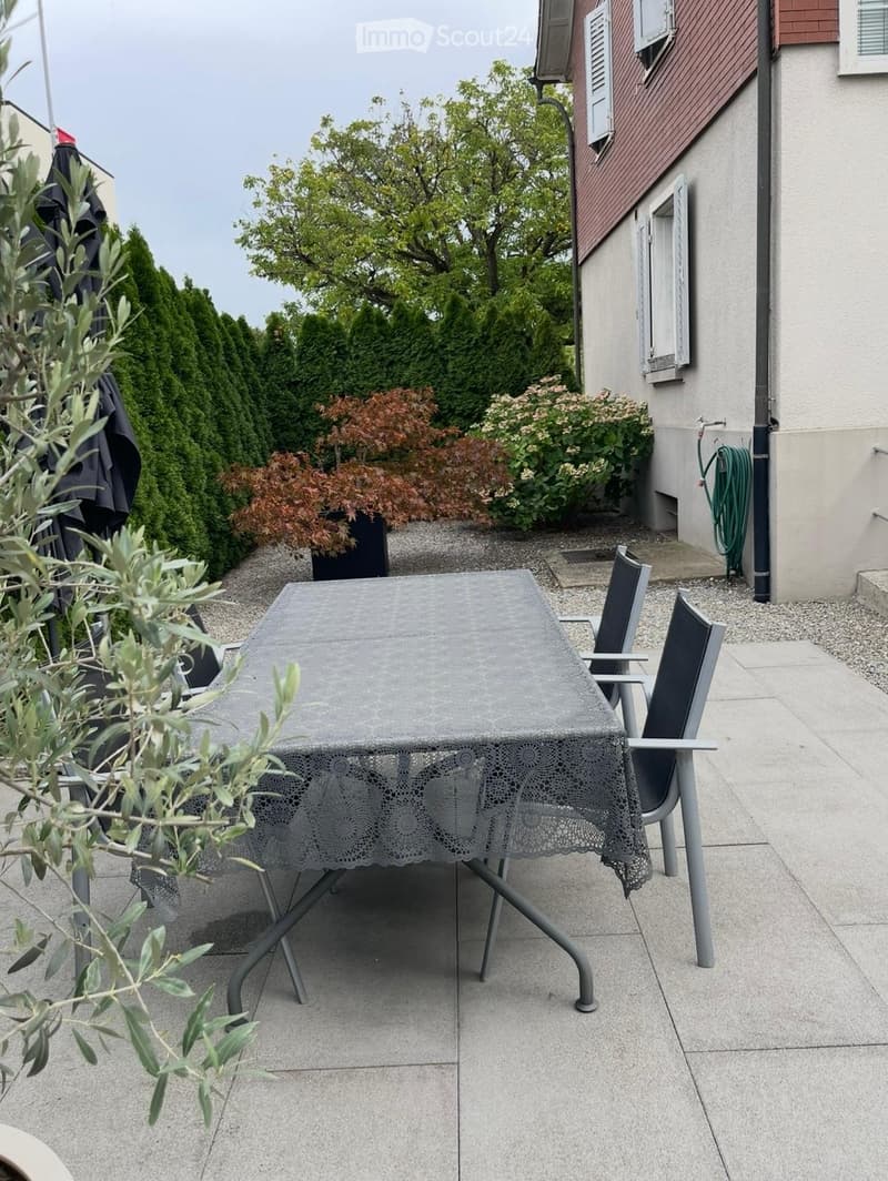 Outdoor patio with a grey table and chairs, surrounded by greenery and a building with shutters.