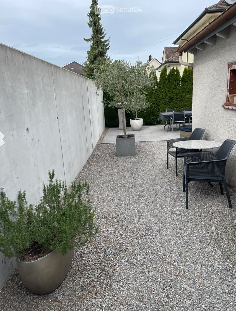 Gravel patio with a small seating area featuring a round table and two black chairs, surrounded by potted plants and a modern concrete wall.