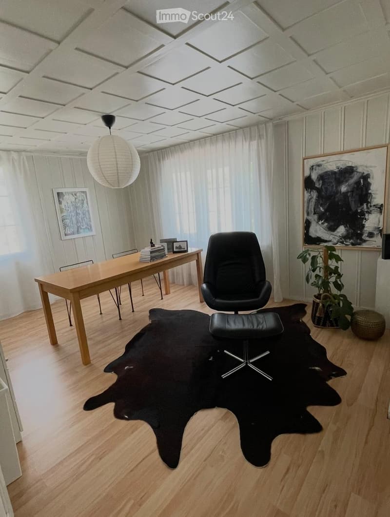 Bright living room with wooden flooring, large window with white curtains, a black leather chair, a wooden table with books, a hanging lamp, and artwork on the walls.