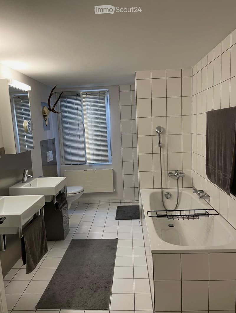Modern bathroom with two sinks, a toilet, a bathtub with a showerhead, and a large window with blinds. The room features tiled walls and floors, a mirror, and decorative antlers on the wall.