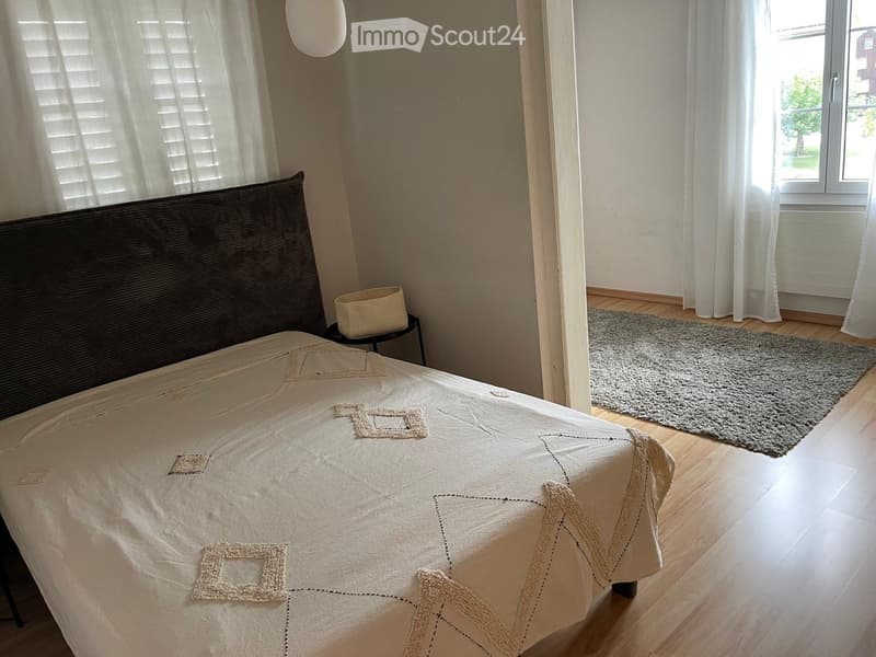 A well-lit bedroom with a large bed, a nightstand, and a gray rug on the wooden floor. The room features white curtains and a window with blinds.