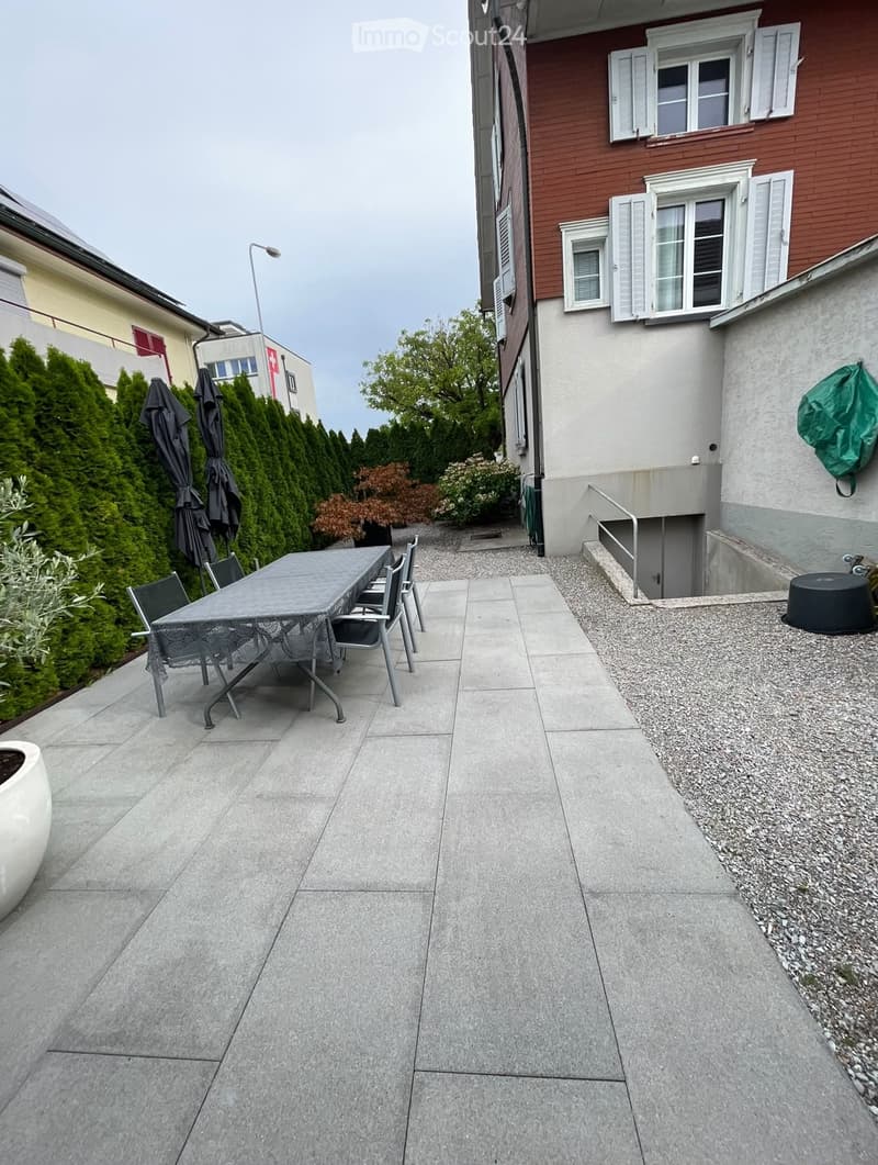 Outdoor patio with a tiled floor, a table with chairs, and surrounding greenery including tall conifers and flowering shrubs. The area is adjacent to a residential building with multiple windows and a staircase leading down.
