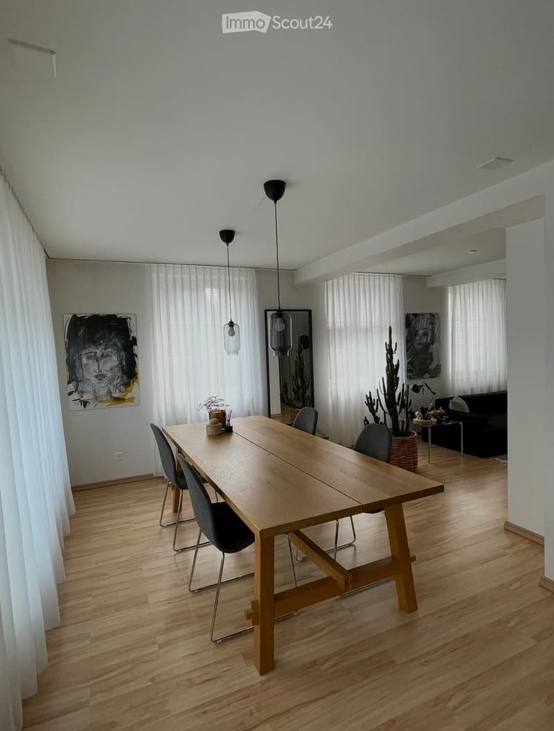 Open-plan dining and living area with wooden flooring, large windows with white curtains, a wooden dining table with four chairs, a black sofa, pendant lights, and decorative artwork.