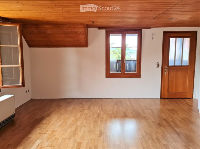 Empty room with wooden flooring, white walls, and large windows, featuring a wooden door and a slanted wooden ceiling.