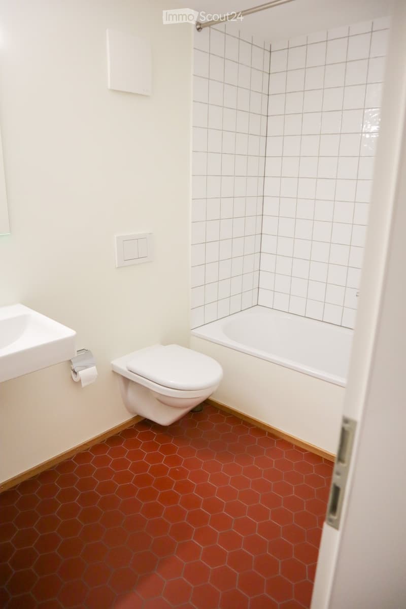Modern bathroom with white tiled walls, hexagonal red tiles on the floor, a toilet, a bathtub, and a sink.