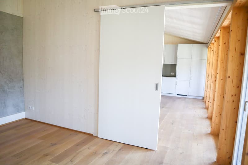 Empty hallway with wooden flooring, white walls, and a sliding door leading to a kitchen area with white cabinets and a countertop.
