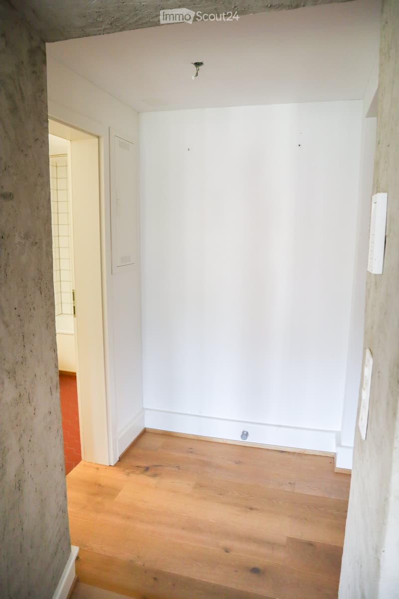 Empty room with wooden flooring and white walls, featuring a concrete pillar and a doorway leading to another room.