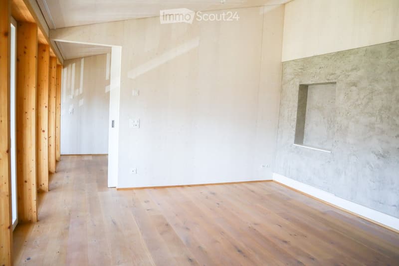 Empty room with light wooden flooring, white walls, and a concrete accent wall. The space features exposed wooden beams and electrical outlets.