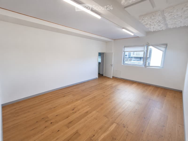 Empty room with light wooden flooring, white walls, and a large window with blinds. The room has a door leading to another space and modern lighting fixtures on the ceiling.