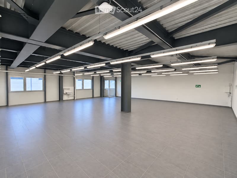 Empty room with tiled floor, white walls, exposed ceiling beams, and fluorescent lighting. Features include a sink, windows, and a central pillar.