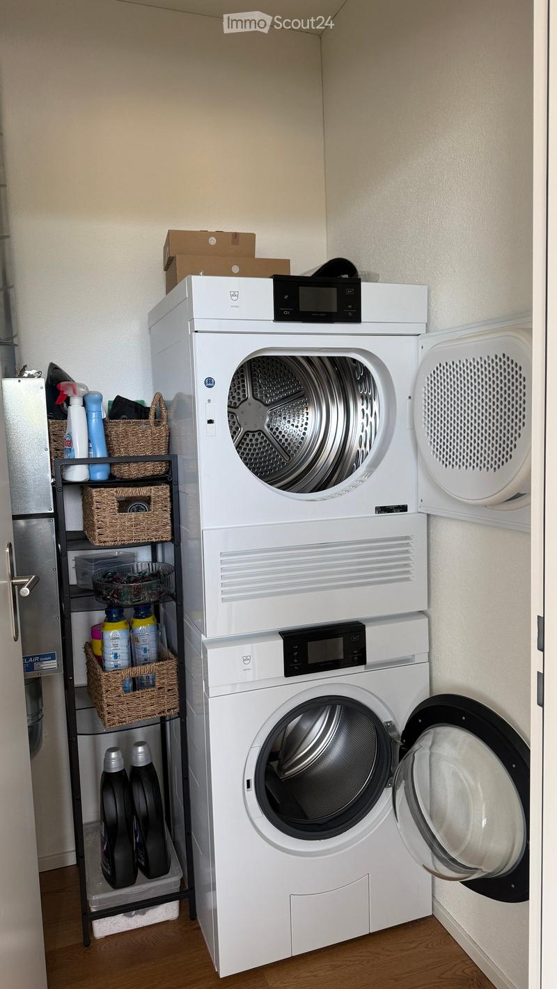 A compact laundry area with a stacked washing machine and dryer, a shelf with cleaning supplies and baskets, and a wooden floor.