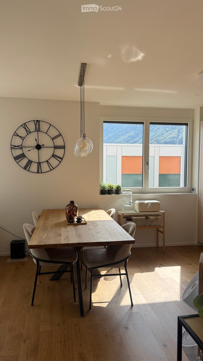 Bright dining area with wooden table, four chairs, decorative vase, wall clock, and a small shelf with items near the window.