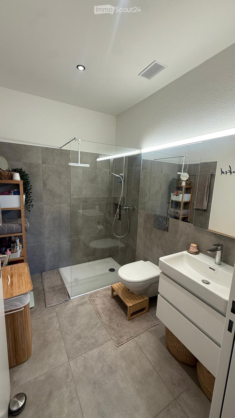 Modern bathroom with a glass-enclosed shower, toilet on a wooden stool, sink with a modern faucet, and a wooden shelf with towels and toiletries.