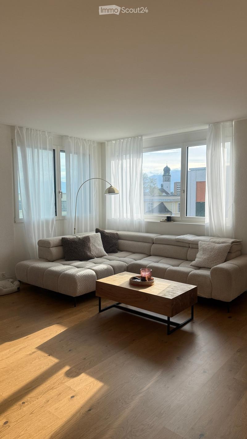 Modern living room with a large sectional sofa, wooden coffee table, floor lamp, and large windows with white curtains. The room features light wooden flooring and a view of a cityscape with a church spire.