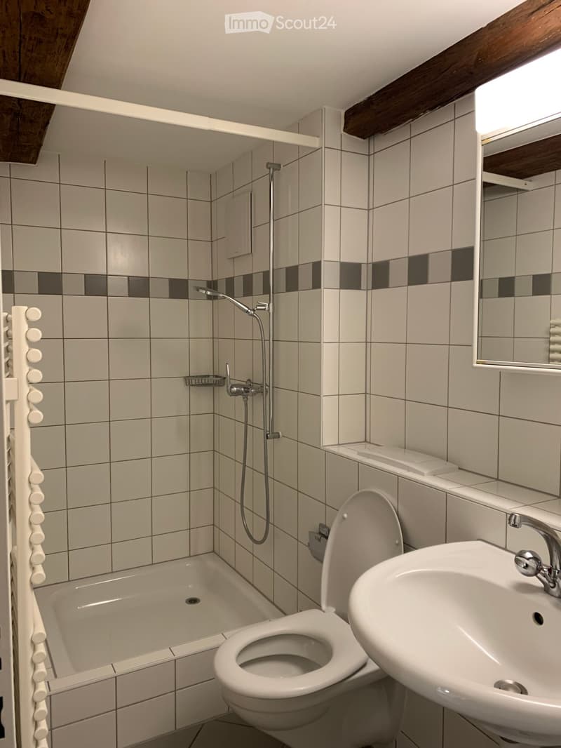 Modern bathroom with white tiles, bathtub, toilet, sink, and showerhead. Features a large mirror and a window with wooden frame.
