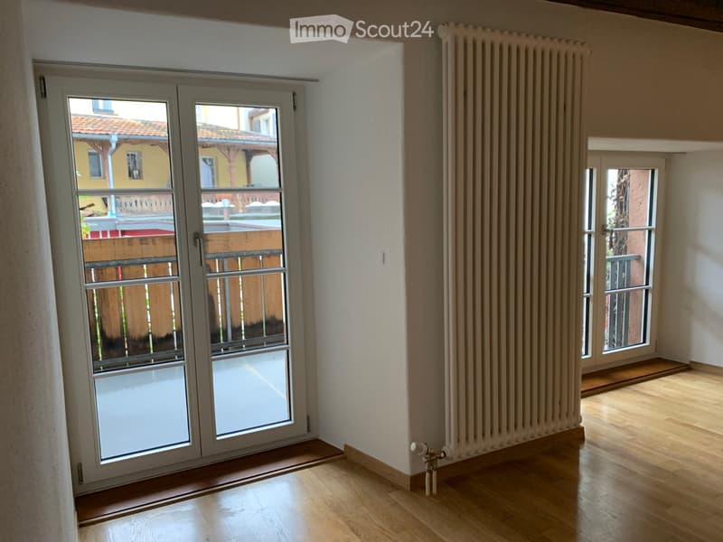 Empty room with wooden flooring, large sliding glass door leading to a balcony, radiator on the wall, and a small window with a view of the street.