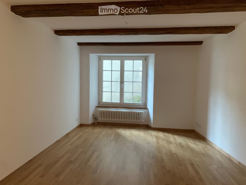 Empty room with wooden flooring, white walls, exposed wooden beams on the ceiling, and a single window with a radiator below it.