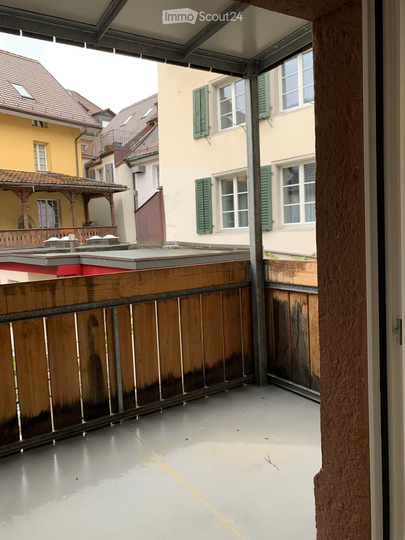 Balcony with wooden railing, concrete floor, overlooking a courtyard with neighboring buildings and green shutters.