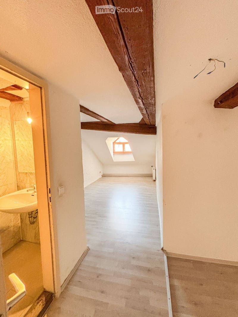 Empty attic space with exposed wooden beams, white walls, and a skylight window. The floor is light-colored and the space is clean and unfurnished.