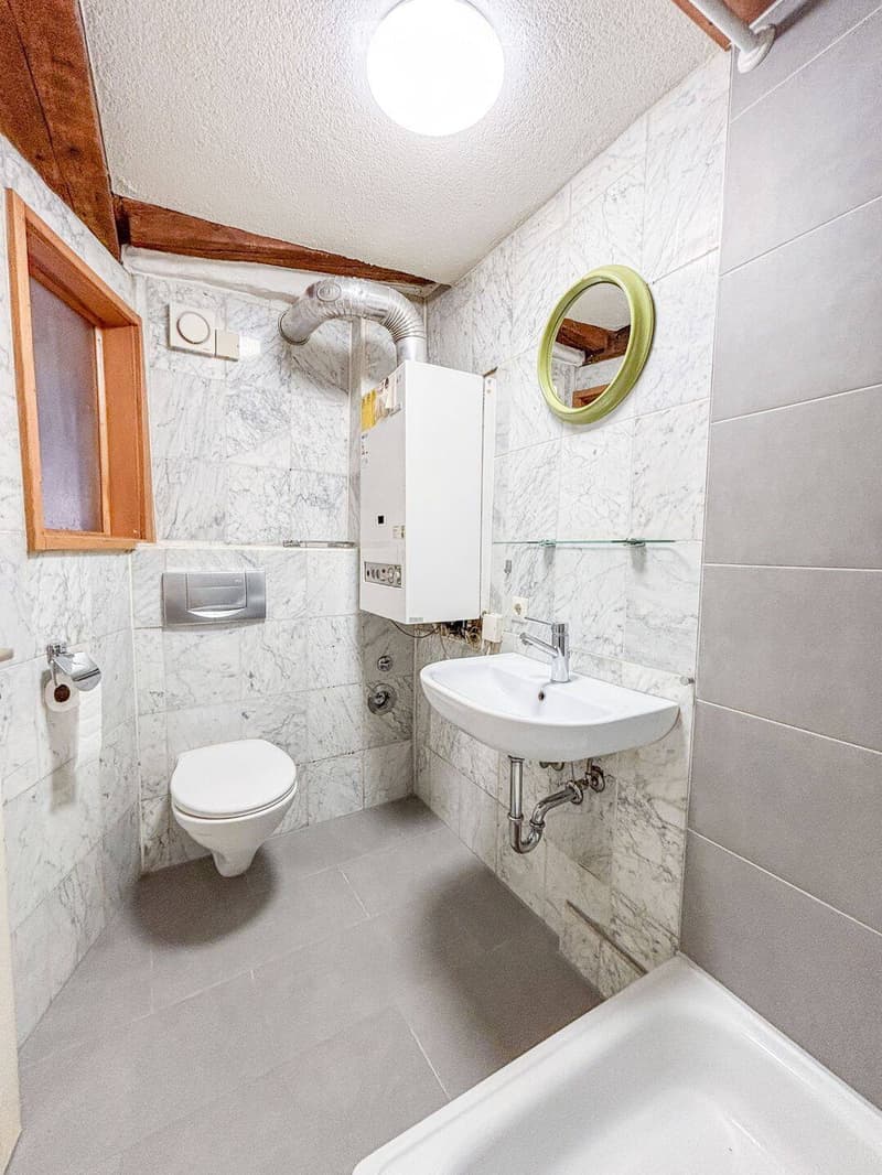 Modern bathroom with white marble tiles, a toilet, a sink with a chrome faucet, a bathtub, a round mirror with a green frame, and a water heater unit.