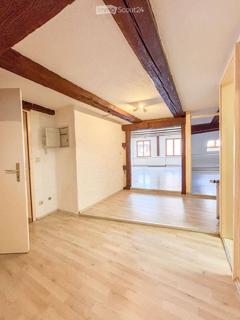 Empty attic room with wooden beams, light-colored wooden flooring, and white walls. Features include exposed wooden beams, a single door, and a small window in the adjacent room.