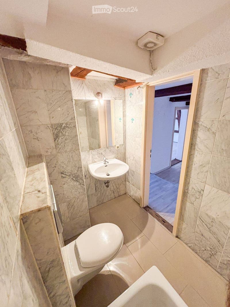 Bathroom with marble walls and floor, featuring a sink, toilet, bathtub, and mirror. An open doorway leads to another room.