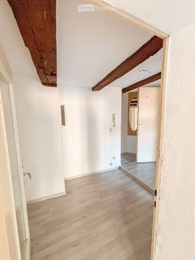 Empty room with wooden flooring, white walls, and exposed wooden beams on the ceiling. A doorway leads to another room with a visible mirror and closet.