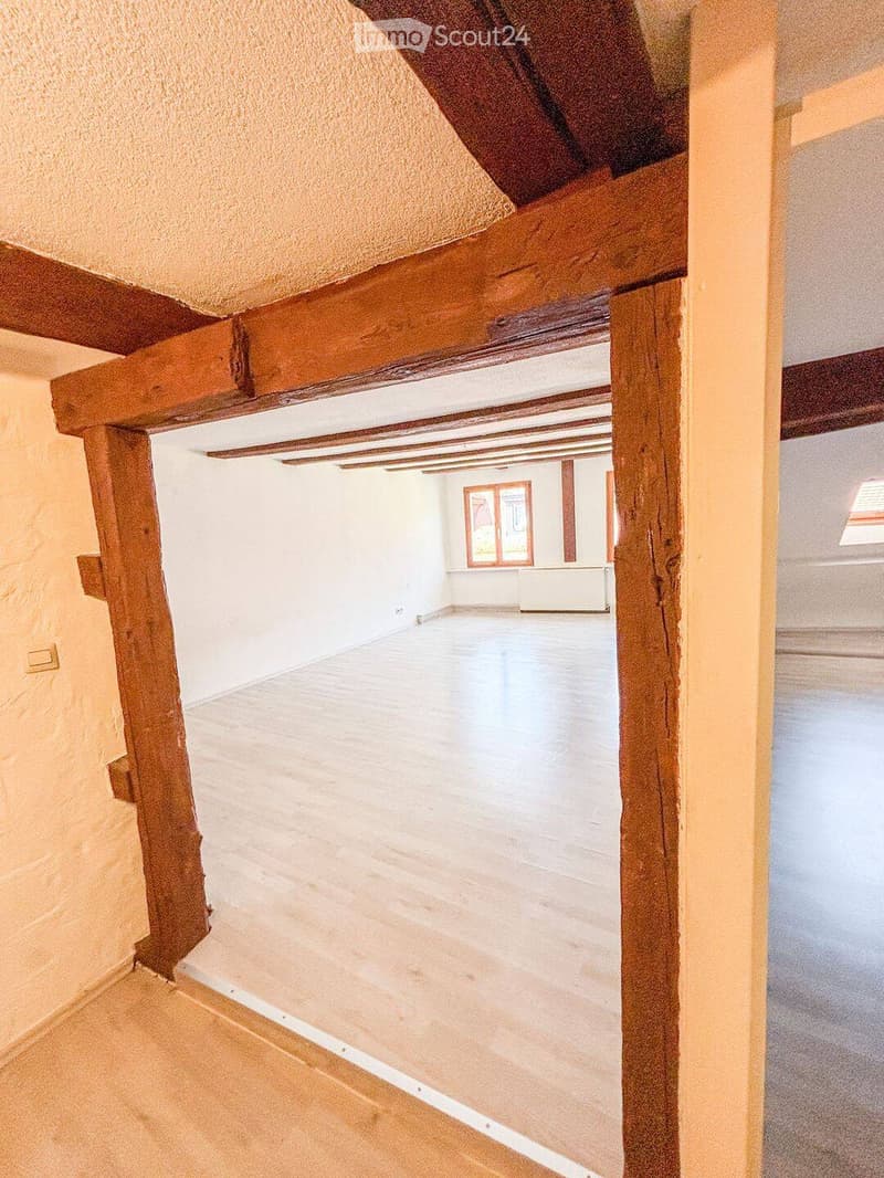 Empty room with wooden beams and white walls, visible through an open doorway. The room has a smooth, light-colored floor and a window with red frames in the background.