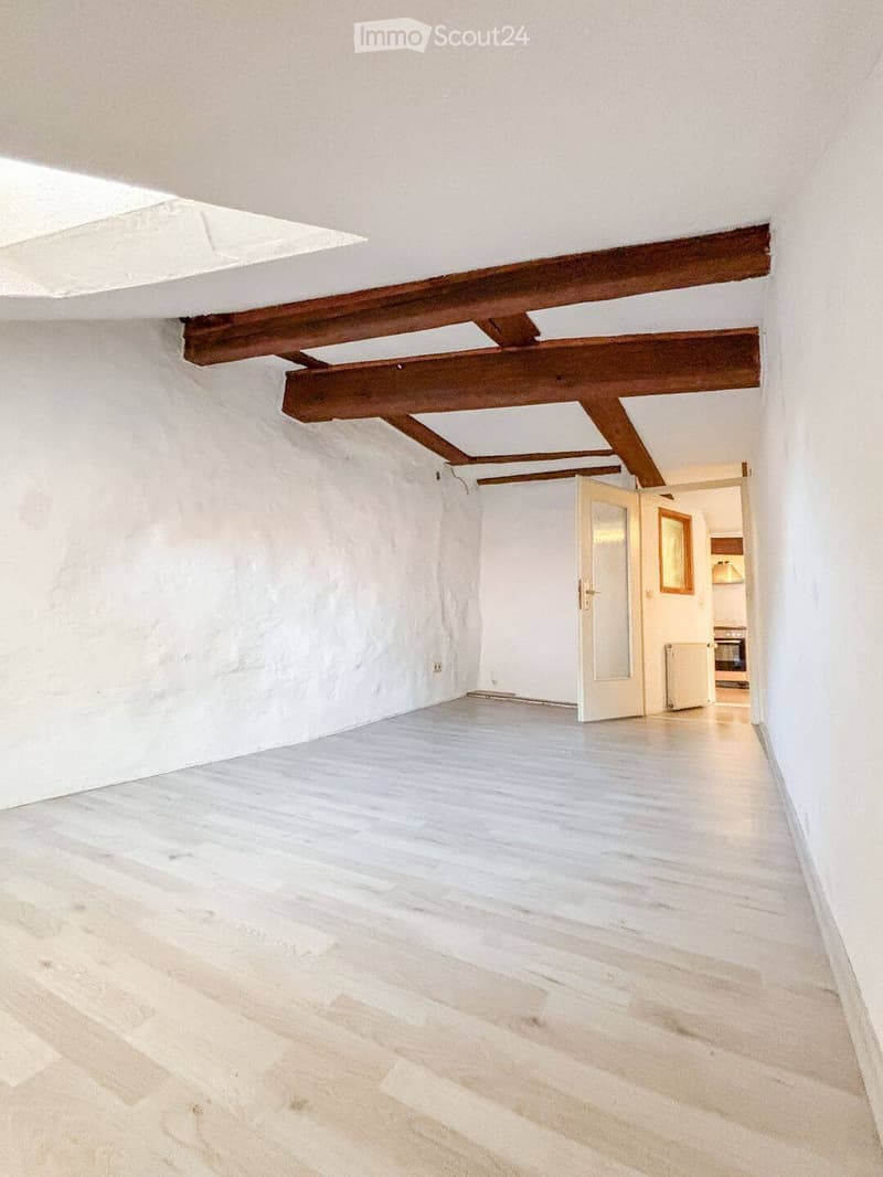 Empty room with white walls, wooden floor, and exposed wooden beams on the ceiling. A white door leads to another room.