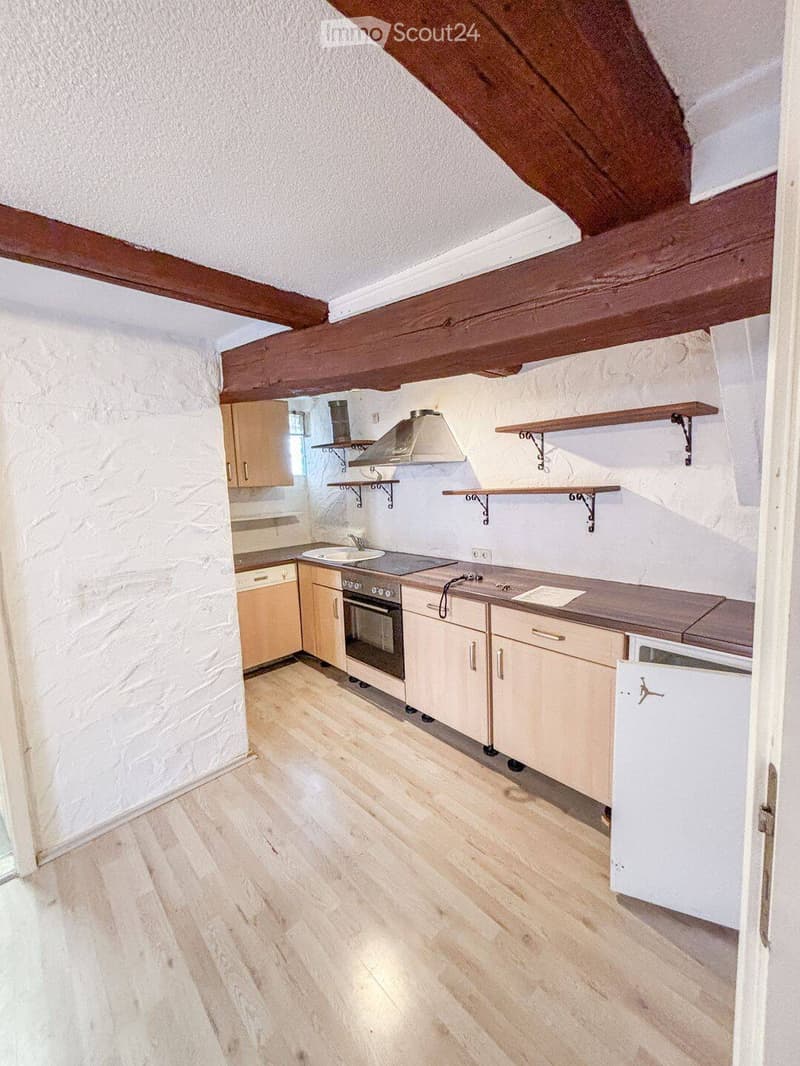 Bright kitchen with wooden flooring, white stone walls, and exposed wooden beams. Features include a sink, oven, cabinets, and wooden shelves.