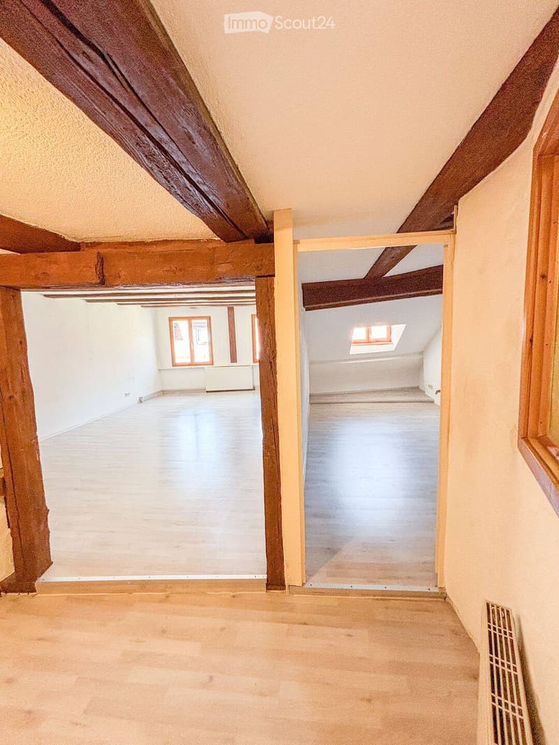 Empty attic space with wooden beams, exposed ceiling, and two rooms visible through an open doorway. The rooms have white walls, concrete floors, and windows allowing natural light.