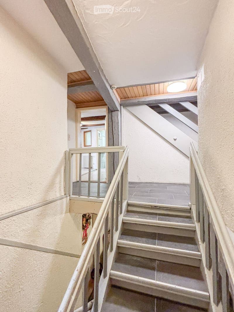 Staircase with wooden handrails and gray steps, leading to an open area with visible ceiling beams and a light fixture.