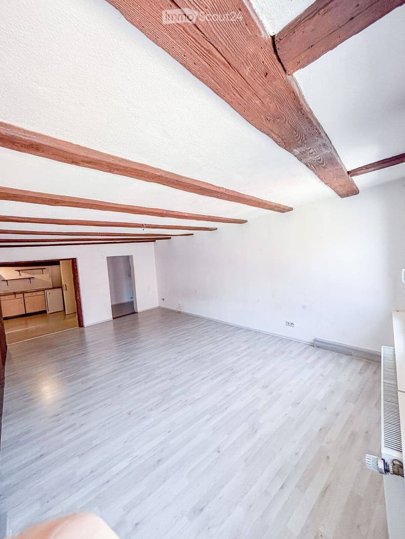 Empty room with exposed wooden beams on the ceiling, light-colored wooden flooring, and white walls. The room has two doorways leading to other spaces.