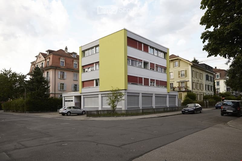 Modern multi-story building with yellow and red facade, surrounded by parked cars and neighboring houses.