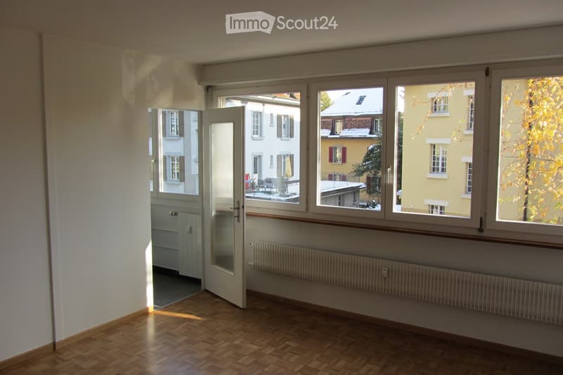Empty room with wooden flooring, large windows, radiator, and an open door leading to a balcony area.