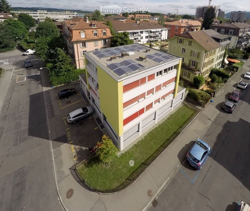 A modern multi-story building with solar panels on the roof, surrounded by parked cars and greenery.