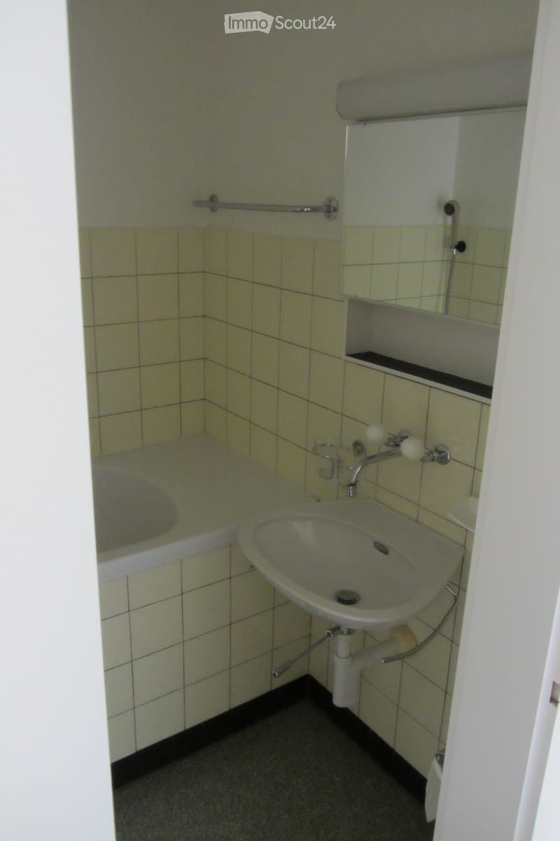 Small bathroom with a sink, bathtub, and mirror. Light-colored tiles on walls and floor, with a towel rack above the bathtub.