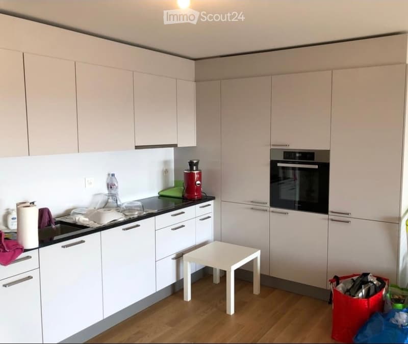 Modern kitchen with white cabinets, black countertops, and a small white stool. Features an oven, coffee maker, sink, and various small items on the counter.
