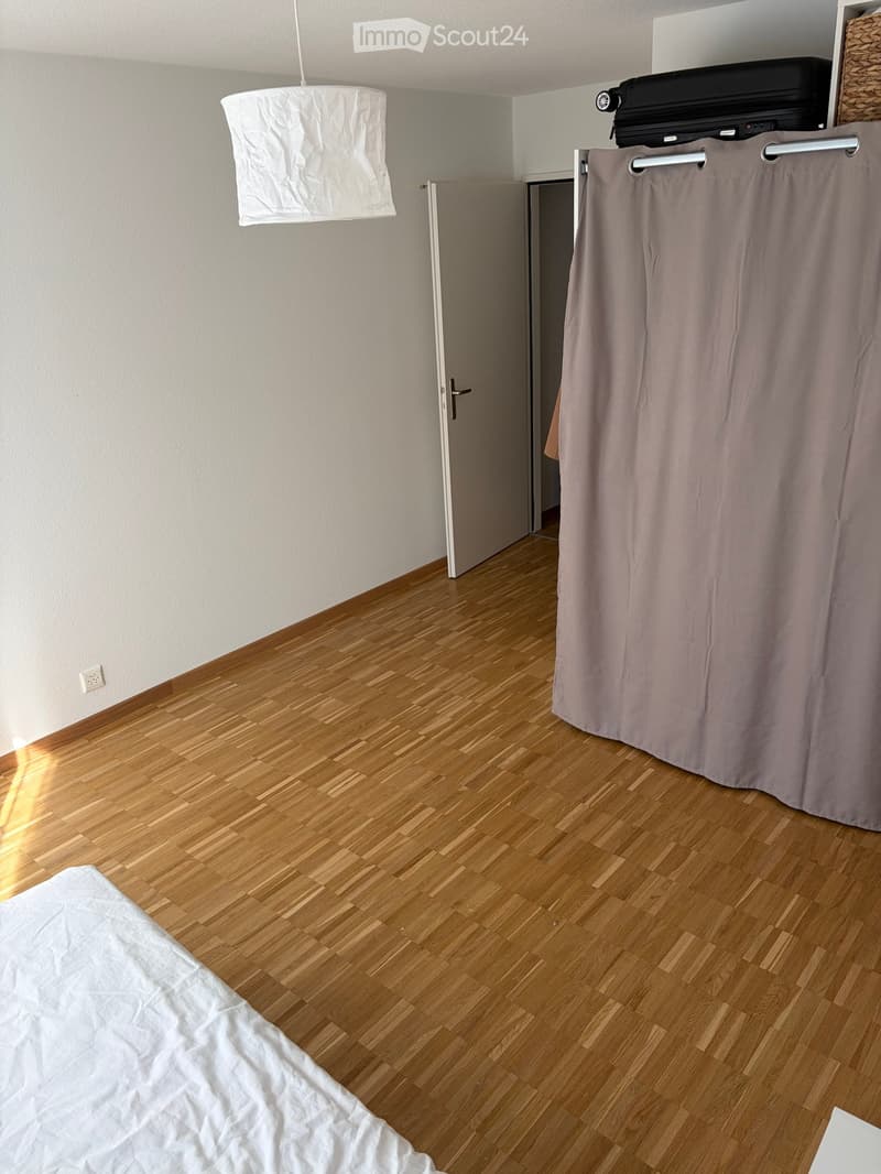 A furnished room with wooden flooring, a bed with white sheets, a curtain, and a suitcase on top of a shelf.