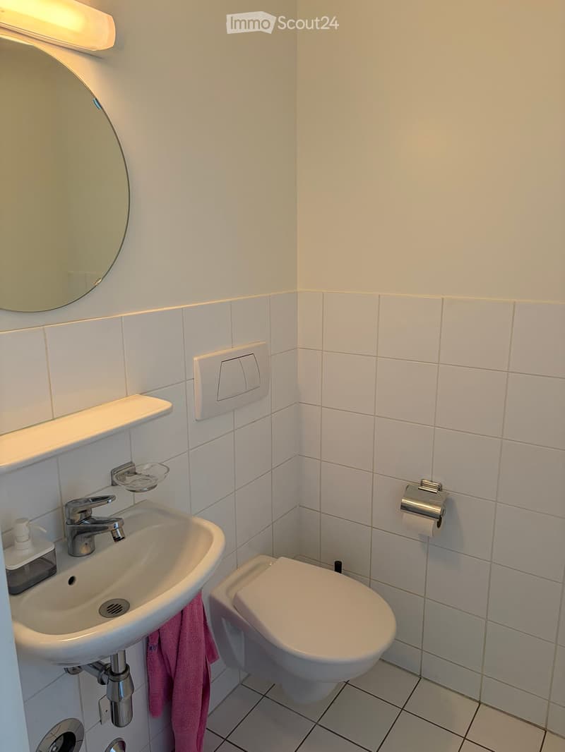Modern bathroom with white tiles, a sink with a modern faucet, a round mirror, a toilet, and a towel hanging on the side of the sink.