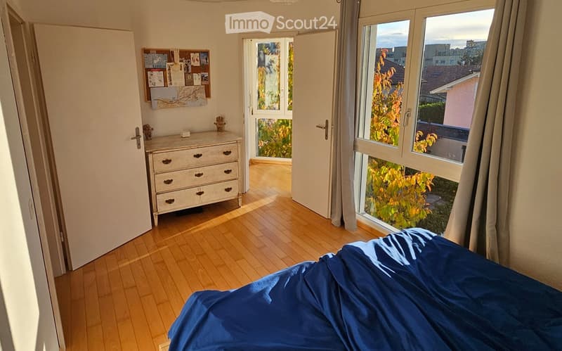 A well-lit bedroom with wooden flooring, a blue bed covered with a duvet, a dresser with drawers, and a large window with a view of the garden.