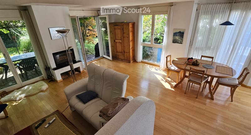 Open-plan living and dining area with wooden flooring, large windows, a fireplace, a sofa, a coffee table, a dining table with chairs, and a floor lamp.