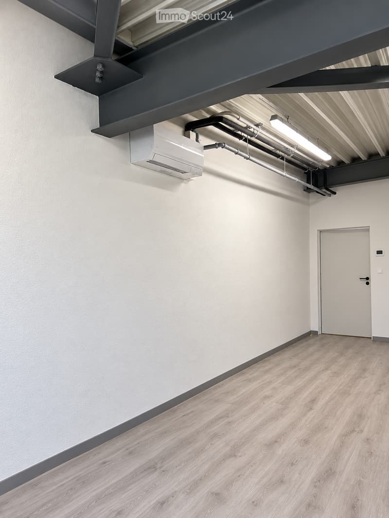 Empty room with light-colored wooden flooring and white walls, featuring a white door and exposed ceiling with lighting fixtures and air conditioning unit.